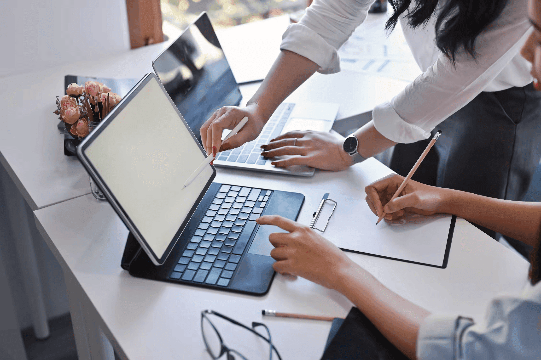Two people working at a desk with a laptop and tablet, one taking notes on a clipboard