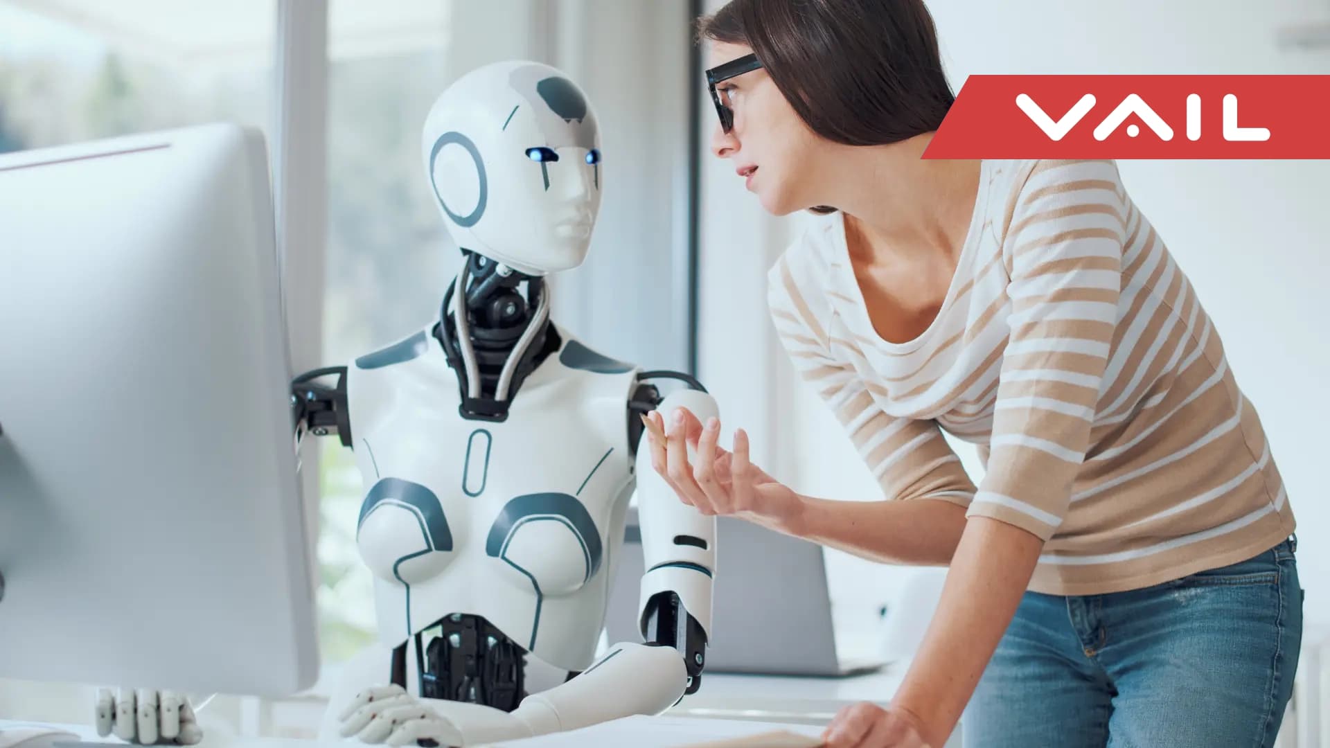 A robot sits at a computer while a woman stands beside it, speaking over its shoulder.