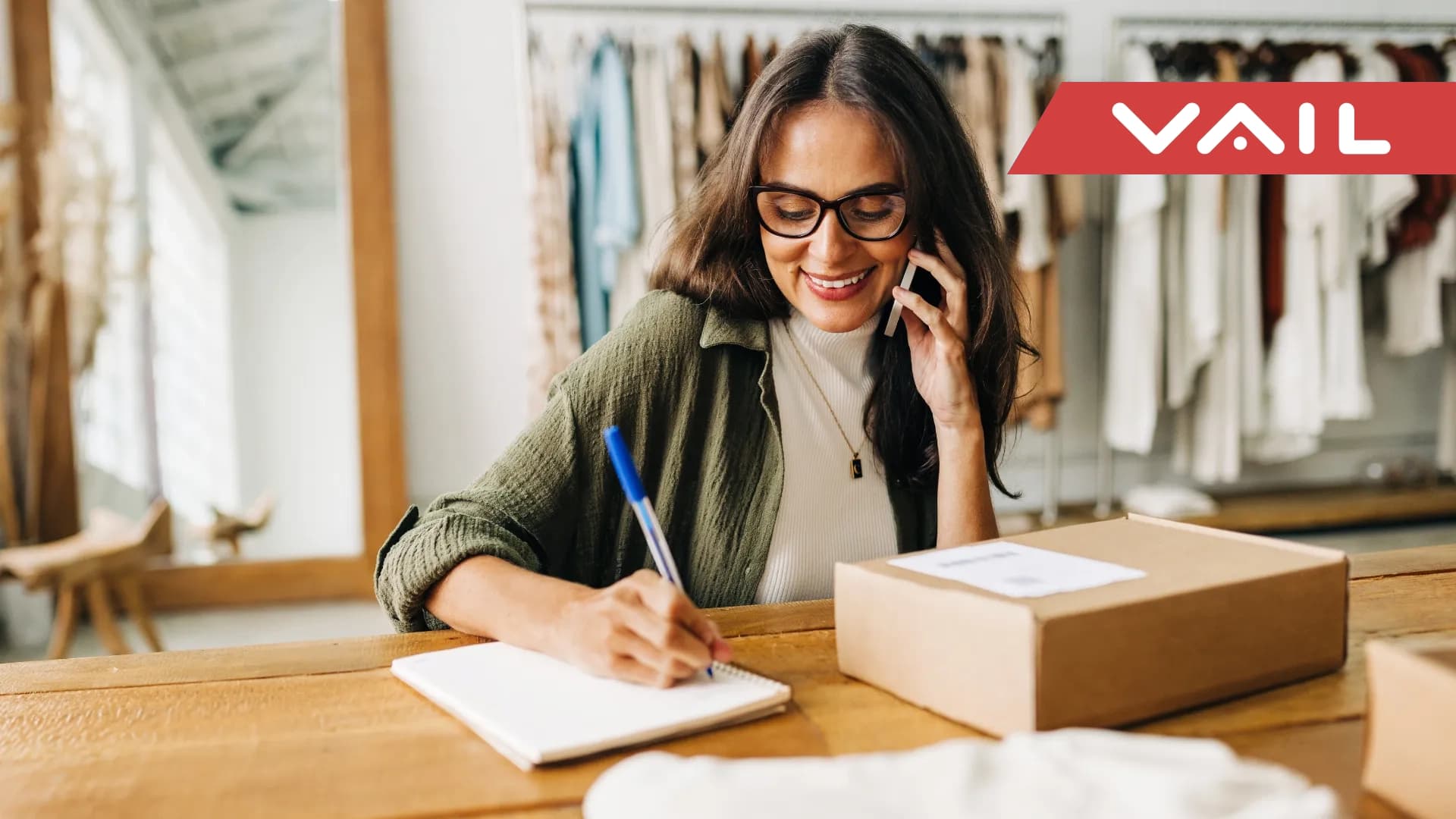 A women in a clothing boutique sits at a desk smiling on the phone while writing in a notebook.