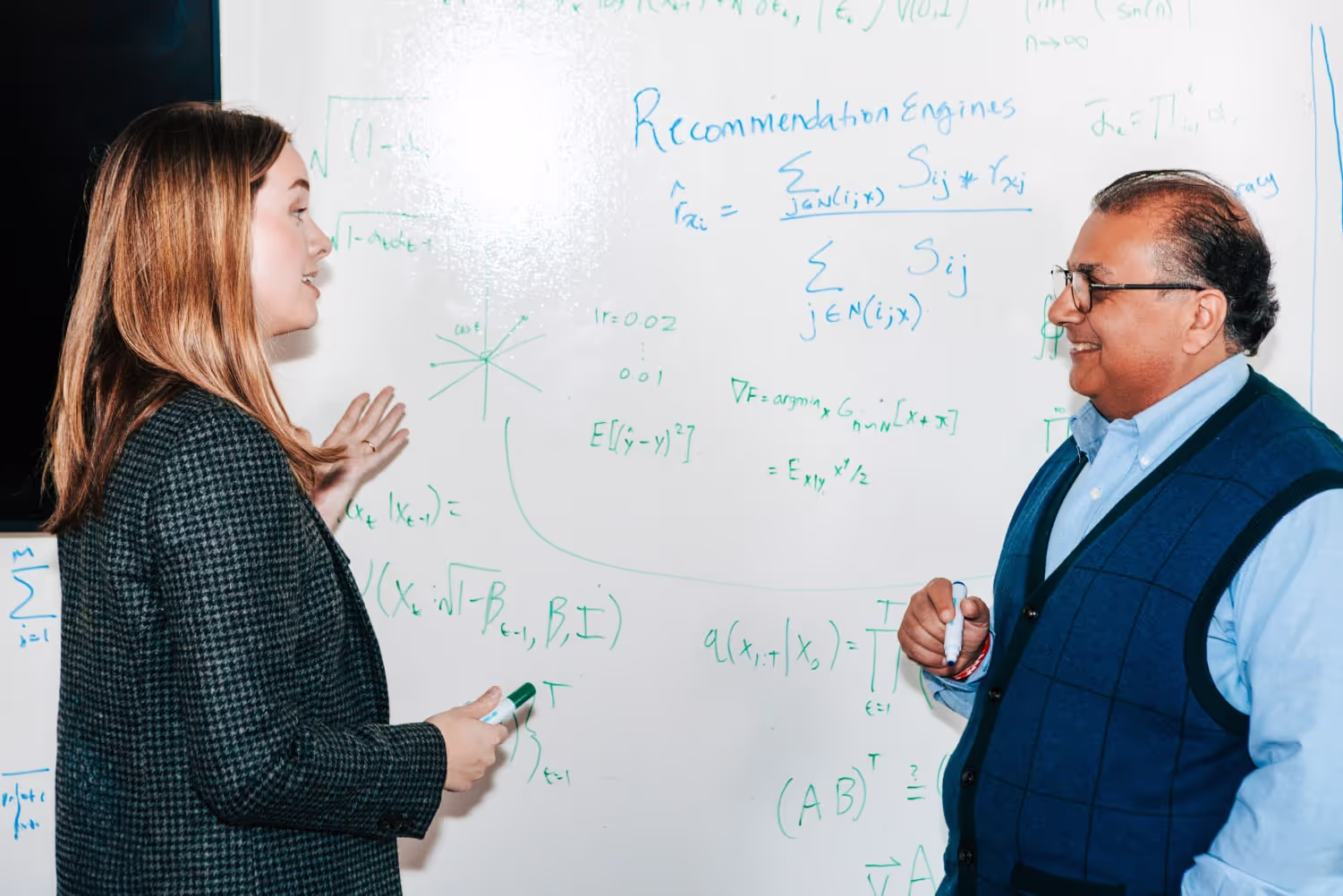 A woman and man with markers talk in front of a whiteboard covered in formulas